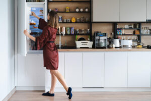 mulher com fome procurando comida na geladeira em casa mas nao tem muito la moveis de cozinha brancos em casa usam roupao de seda vermelho